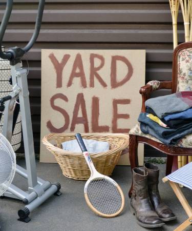 cardboard yard sale sign with an exercise bike, tennis racket, chair, boots, and clothing surrounding it