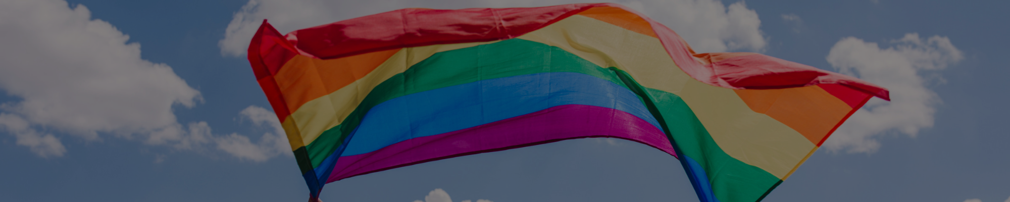 Rainbow flag with blue skies and clouds behind it.