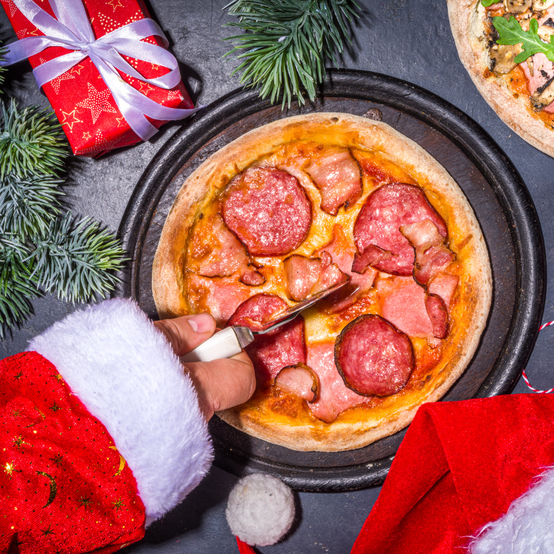 image of someone in a Santa suit cutting pizza with Christmas decorations next to it and a Santa hat