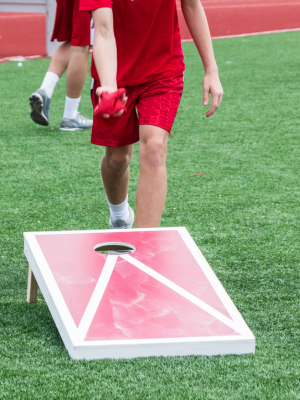 Image of two people playing cornhole. It is not showing their face, you can see one person throwing, another person in the background, and a cornhole board to the left of the person throwing the beanbag.