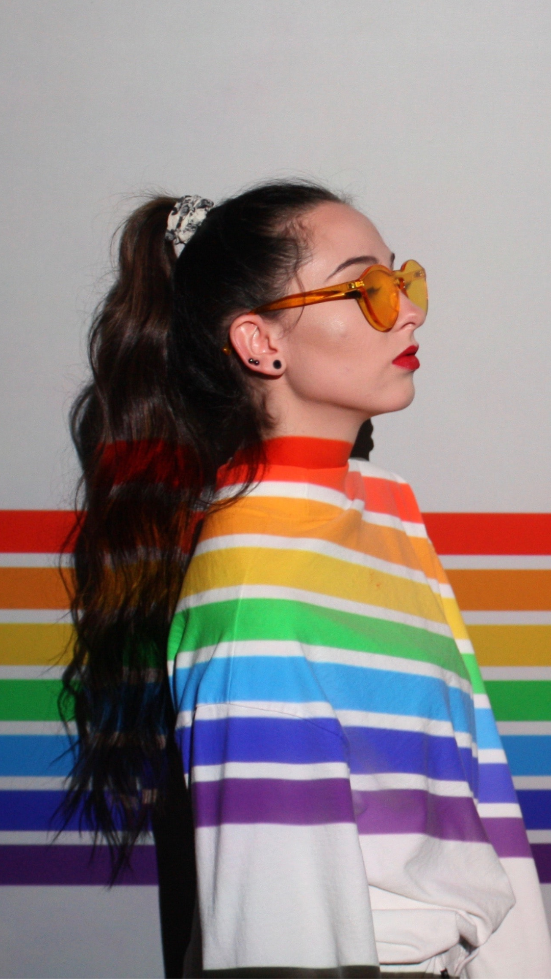 Image of a woman wearing glasses, standing in front of a wall, with the pride colors painted horizontally on the wall and the paint going over the woman's shirt.