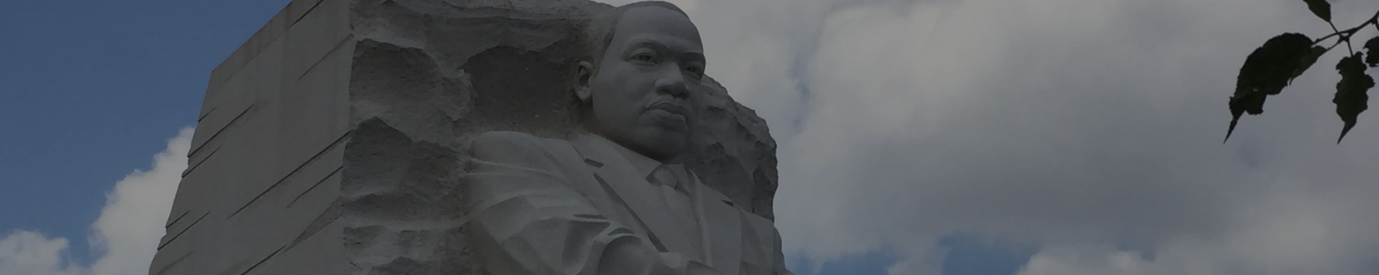Image of a statue of MLK JR with a tree on one side and clouds in the background.